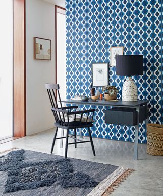 A home office with feature wall papered in a geometric pattern, with tiled floor and rug, desk and armchair.