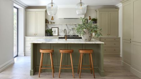 A kitchen with sage green cabinets and a darker green island. A large stone vase with green steps on the island beside a sink, and two glass pendants hanging from the ceiling above. 
