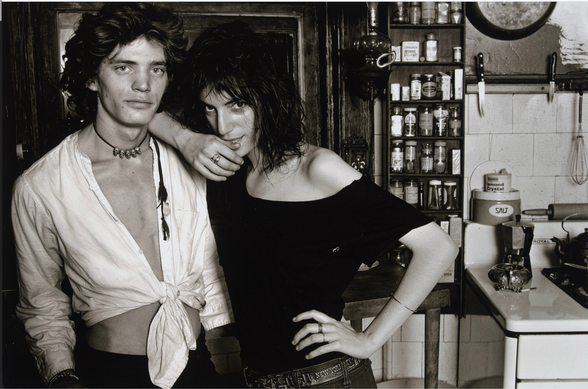 A young man in an open shirt and a woman in a black top stand close together in a cluttered vintage kitchen, both looking directly at the camera.