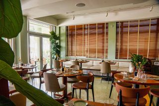 An Art Deco and mid-century modern-inspired cafe and restaurant features brown leathery seats, round wooden tables, wooden blinds, and a green color palette spanning walls and floors.