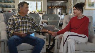 From left to right: Derek sitting in an arm chair and Liz is sitting in an armchair next to him. They are holding hands. 