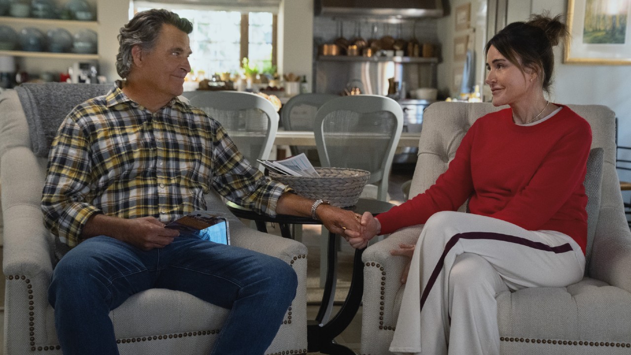 From left to right: Derek sitting in an arm chair and Liz is sitting in an armchair next to him. They are holding hands. 