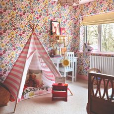 A pink children's room with a play area with a desk, a striped teepee tent and floral wallpaper on the walls