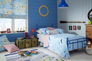 A children's bedroom decorated with a nautical theme with dark blue wood panelled walls, a decorative oar, a decorative porthole, a cast iron blue bed and a storage trunk