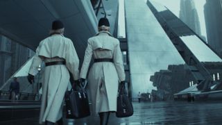 Still from the Star Wars TV show, Andor. Two people, each wearing a white trench coat, black hat, and carrying a briefcase-like bag, are marching towards a towering, sleek-looking building. The weather is miserable and the whole scene is washing out, drab, and gray.