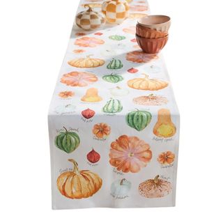 A white table runner with yellow, orange, and green pumpkin illustrations on it and bowls on top