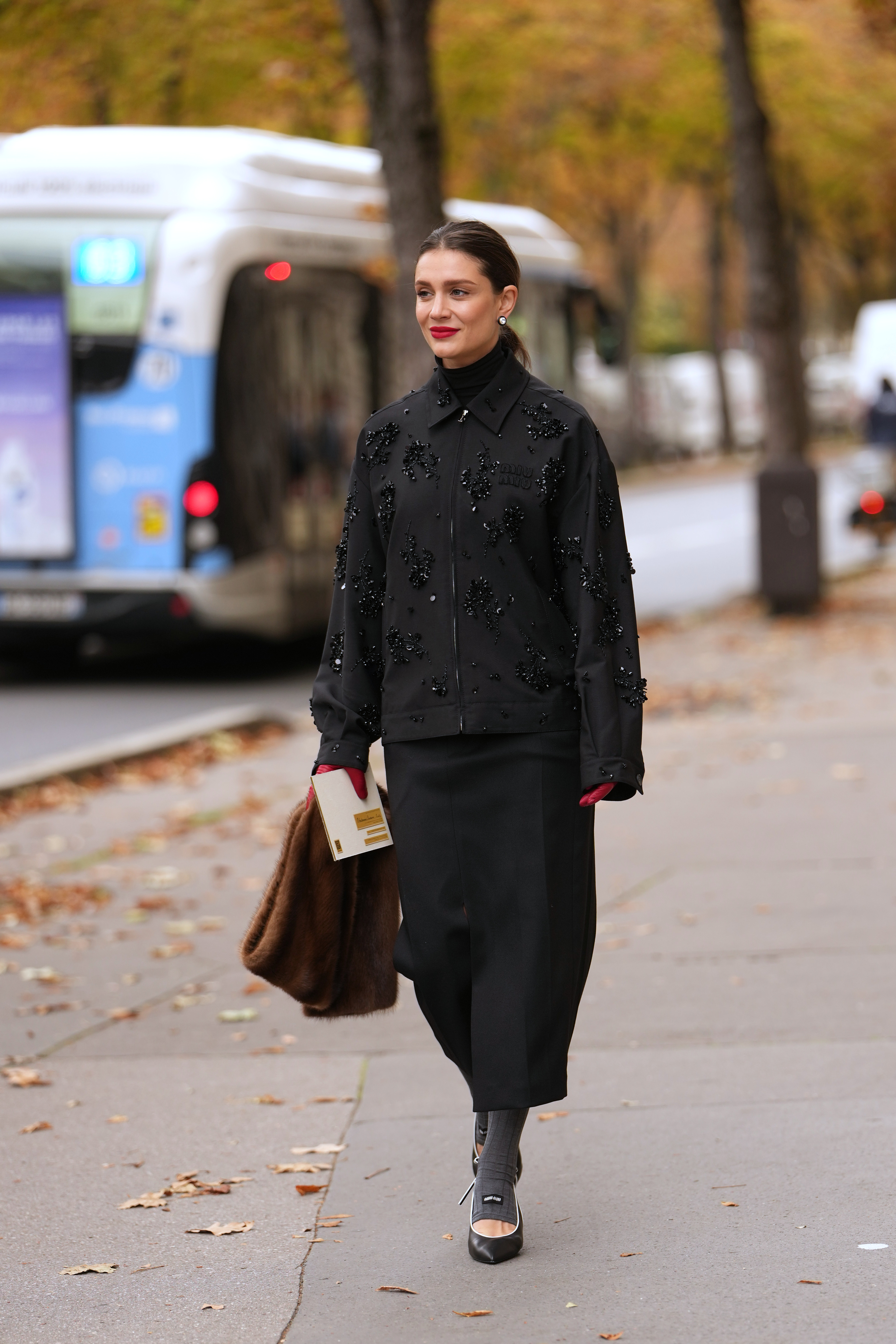 A woman wears a black jacket with sequin details, a black pencil skirt, gray socks, black heels, and a brown fur scarf.