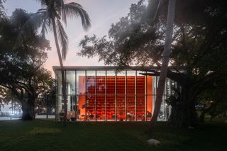 External view of the Wutopia lab builidng, grass verge, tall trees surrounding landscape, glass wall looking into the building, orange shelving unit dominates the room, people sat at some of the table and chairs, dusk sky