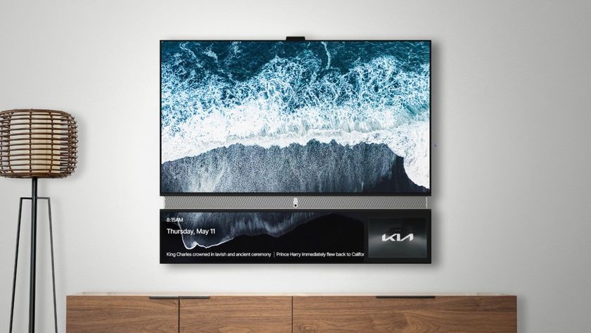 An image of a mounted television displaying an aerial view of waves crashing on a black sand beach, with a secondary lower screen showing the time, date, news headlines about the King&#039;s coronation and Prince Harry, and a KIA logo.