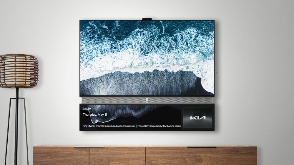 An image of a mounted television displaying an aerial view of waves crashing on a black sand beach, with a secondary lower screen showing the time, date, news headlines about the King's coronation and Prince Harry, and a KIA logo.