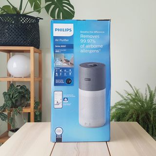 The Philips 3000i Series AC303330 Connected Air Purifier in a blue packaging box on a wooden table in a green room with indoor plants