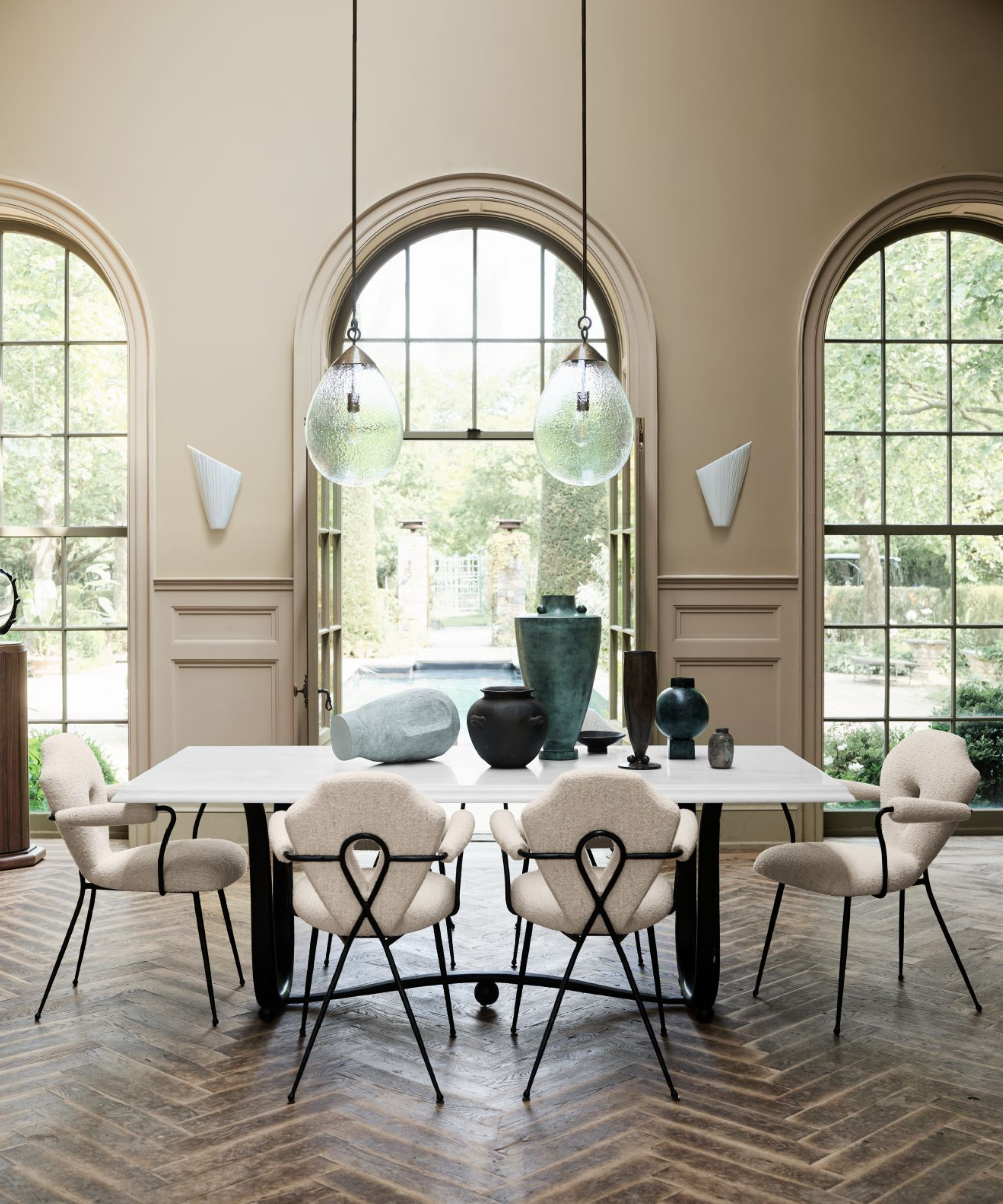 a dining room with four white dining chairs, a marble dining table, various vessels on top of the table, two large hanging pendant lights, and in the background, three large windows
