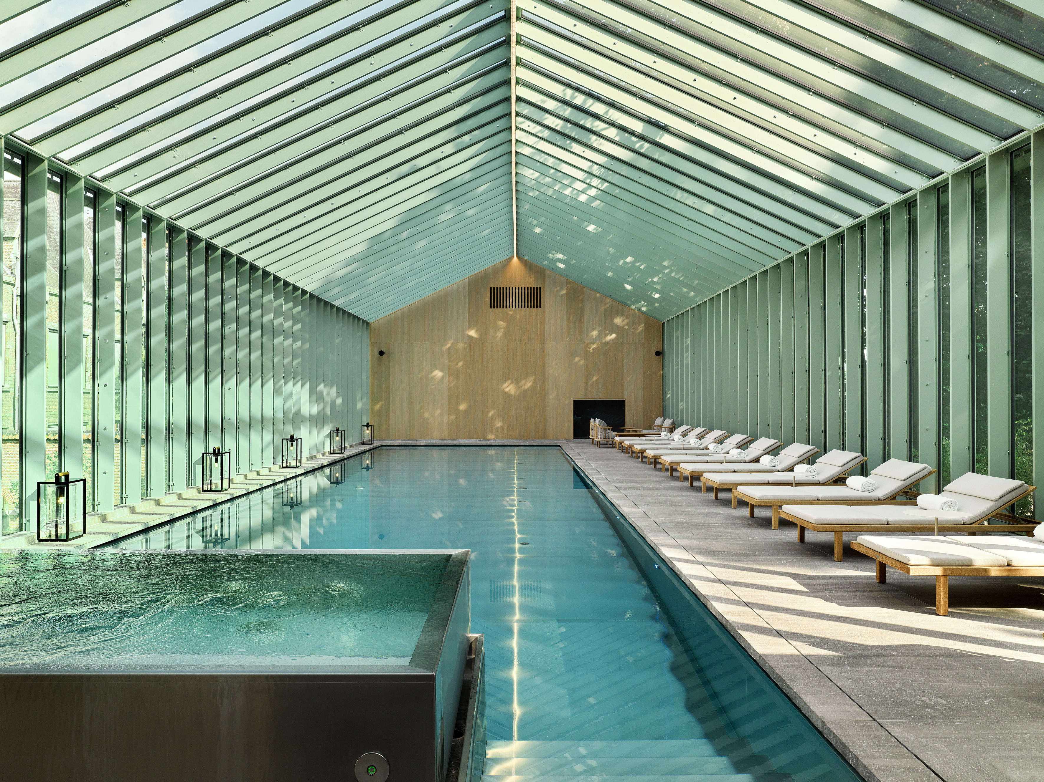 The greenhouse-like, glassy spa pool of a hotel, tucked inside a pavilion, features pale wood ceilings, cream plaster walls, and reclining loungers with structured gray cushions and pale wood backs.