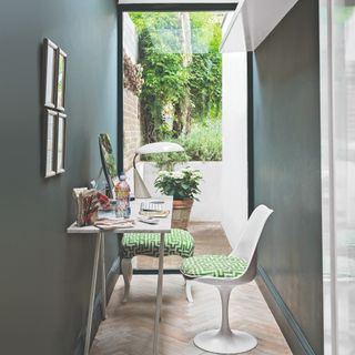 A small narrow home office painted in dark green with a glass wall at the back and a white desk