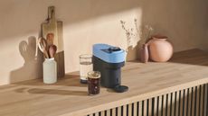 Image of a blue and black Nespresso Vertuo Pop machine on a light wooden countertop with a pink vase, a white utensil holder, and a wooden cutting board also on the counter.
