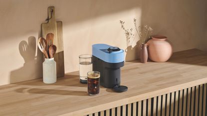 Image of a blue and black Nespresso Vertuo Pop machine on a light wooden countertop with a pink vase, a white utensil holder, and a wooden cutting board also on the counter.