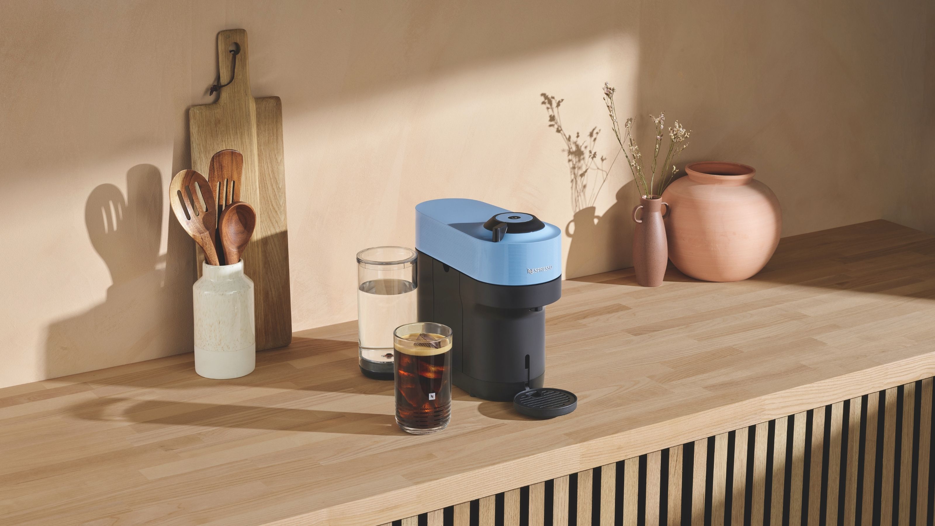 Image of a blue and black Nespresso Vertuo Pop machine on a light wooden countertop with a pink vase, a white utensil holder, and a wooden cutting board also on the counter.