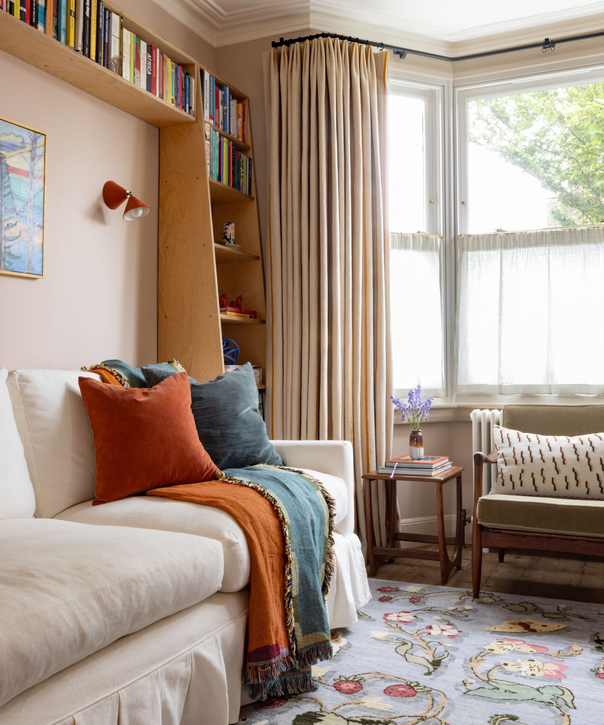 Living room with bay window, peach walls, cream skirted sofa with orange throw, wood bookshelf and midcentury chair