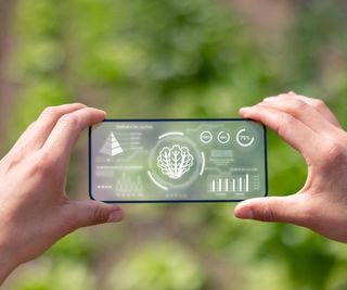 Hands hold up a smartphone. The phone's screen shows an array of data about a plant