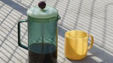 Image of a teal glass French press on a concrete surface with shadows. There is a light yellow mug next to it.