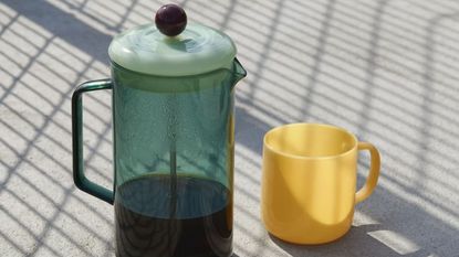 Image of a teal glass French press on a concrete surface with shadows. There is a light yellow mug next to it.