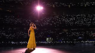 Taylor Swift on stage in a yellow dress for her Eras Tour performance with a purple spotlight on her