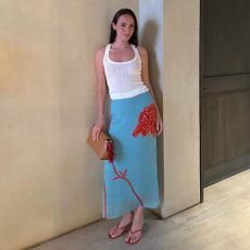 a woman wearing a white tank top, blue and red skirt, red flip-flops, and a tan clutch bag