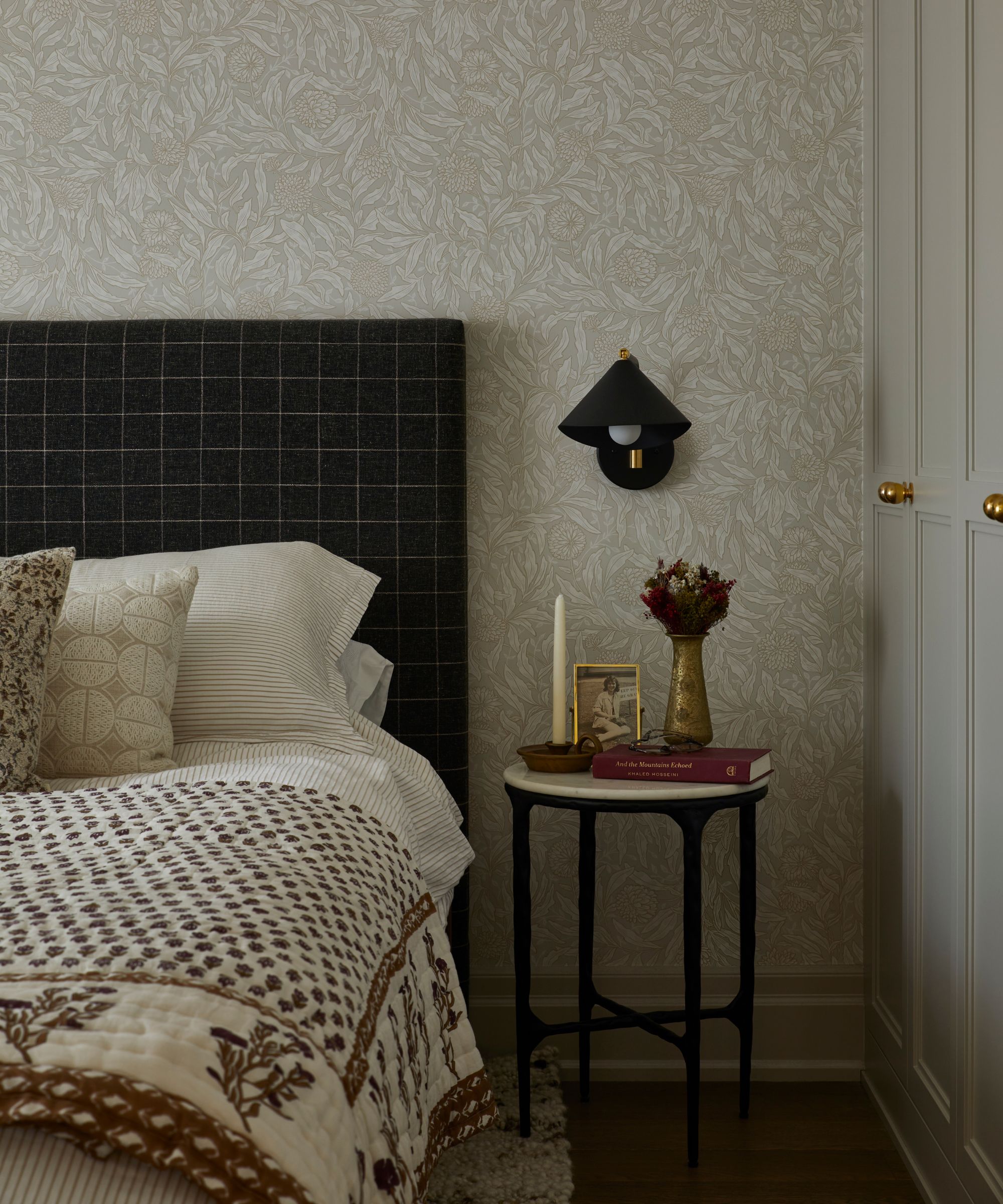 a primary bedroom with neutral beige and white floral wallpaper, beige joinery, a black checked upholstered headboard and a block print floral throw on the bed