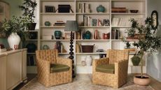 A living room with floor-to-ceiling white bookshelves behind two rattan wicker arm chairs with green cushions.
