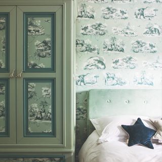 A sage-green bedroom with the walls and panels of the wardrobe covered in a toile de jouy wallpaper