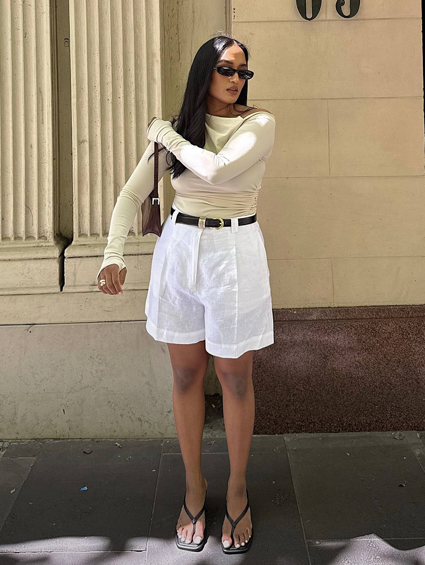 Australian style influencer Div Ravindran wearing black oval sunglasses, a beige long-sleeve stop, small brown leather shoulder bag, high-waisted long linen shorts with a black belt, and black thong flip-flop sandals.