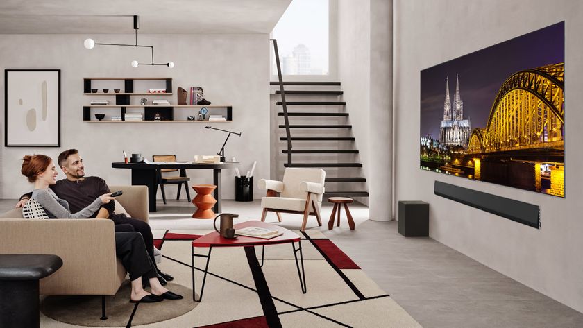 Two people sitting in a modern living room watching a wall-mounted LG G5 OLED TV. The TV is displaying a cityscape at night.