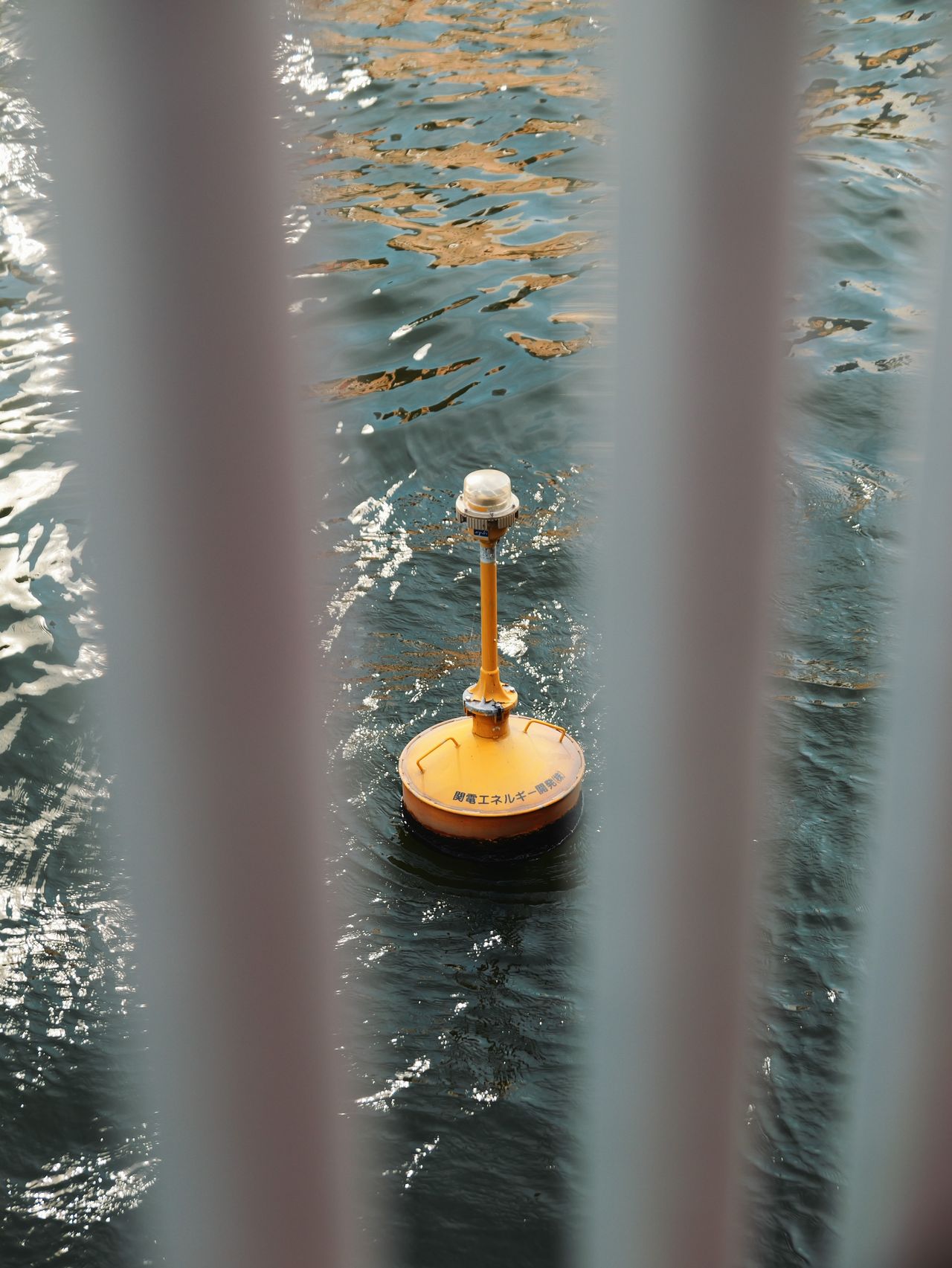 Yellow buoy floating on water, viewed through bridge railings.