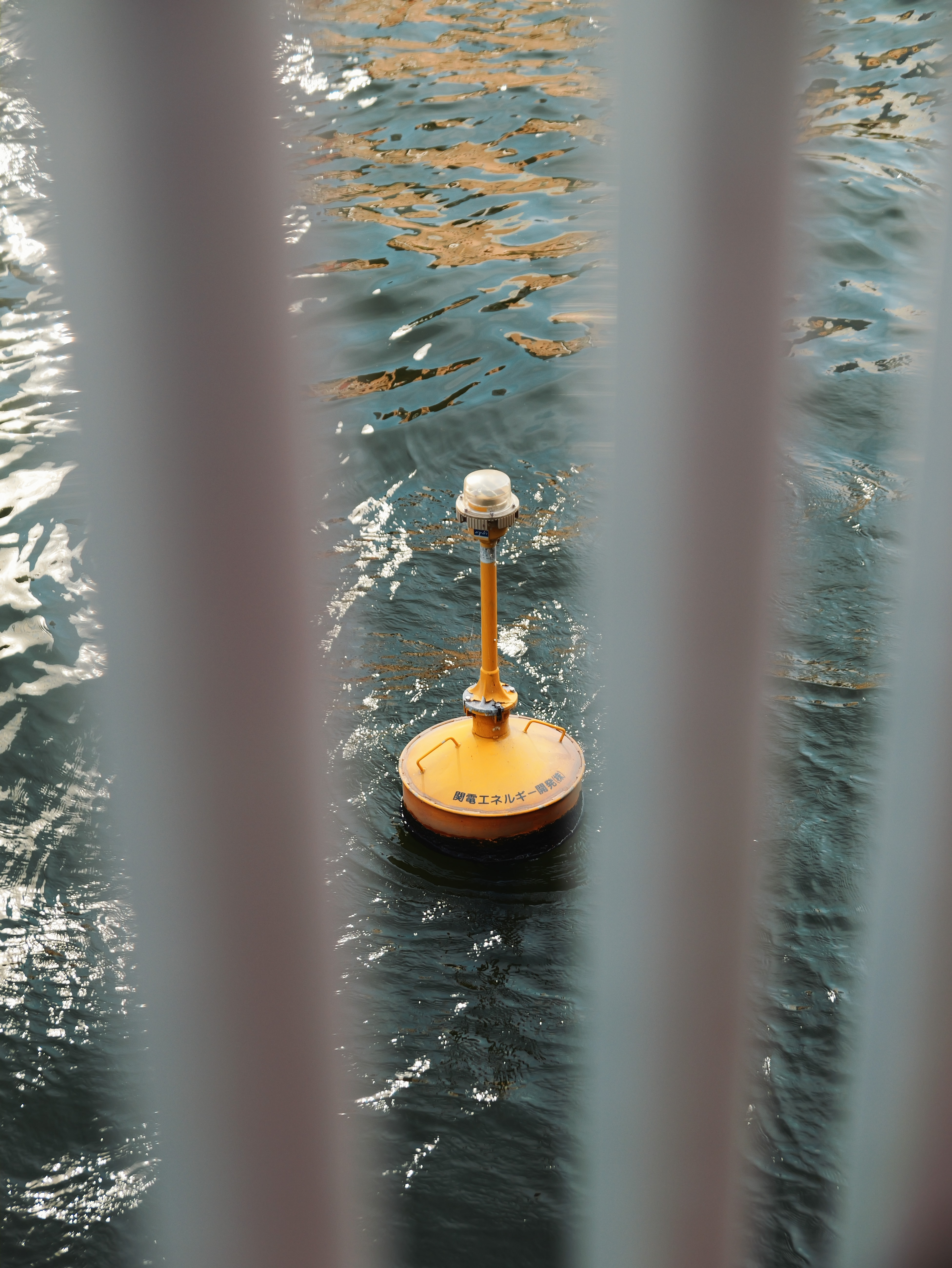 Yellow buoy floating on water, viewed through bridge railings.