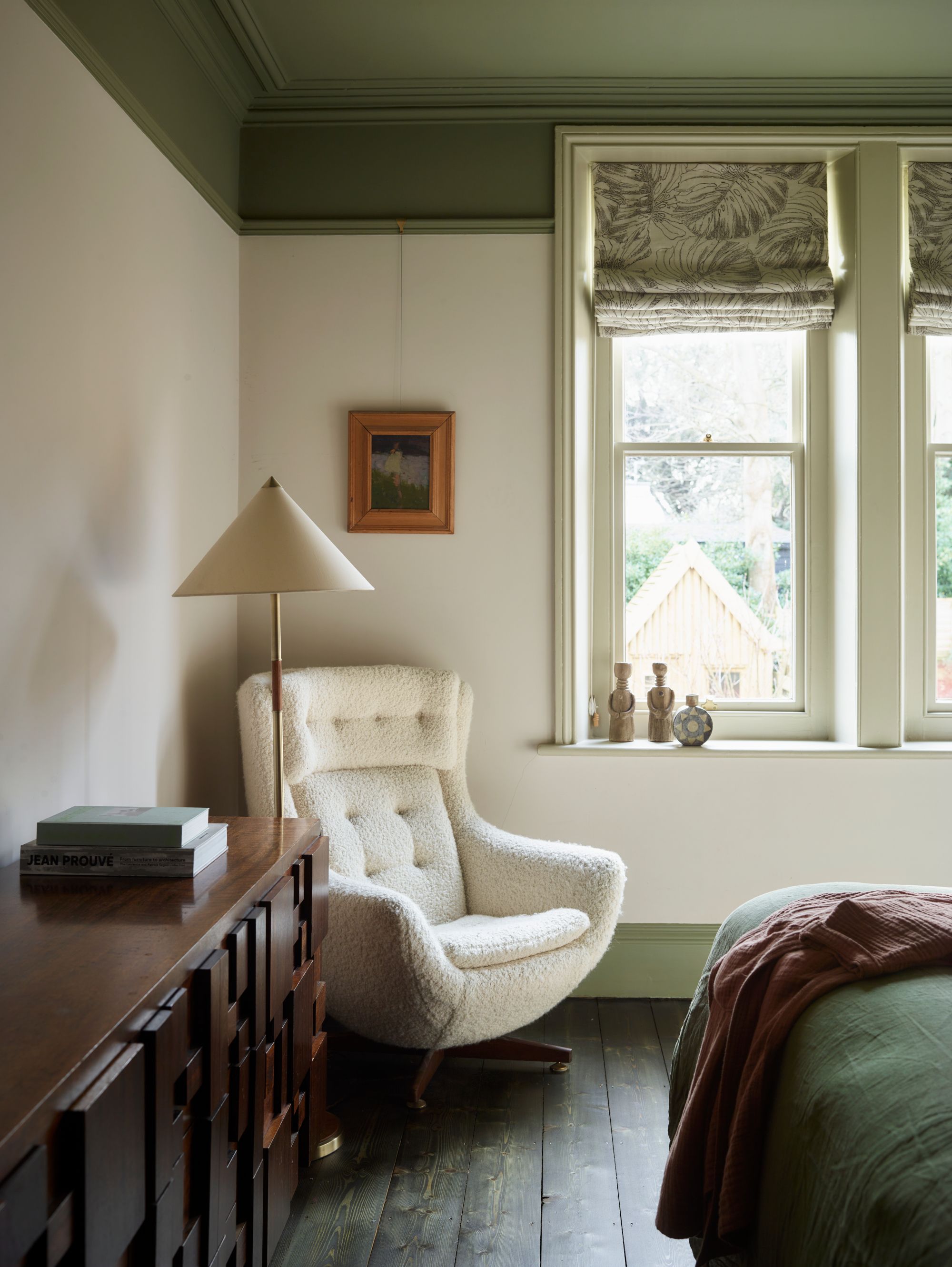 Corner of a bedroom with color-capped green ceiling and white walls, white boucle armchair, cream floor lamp and dark wood sideboard