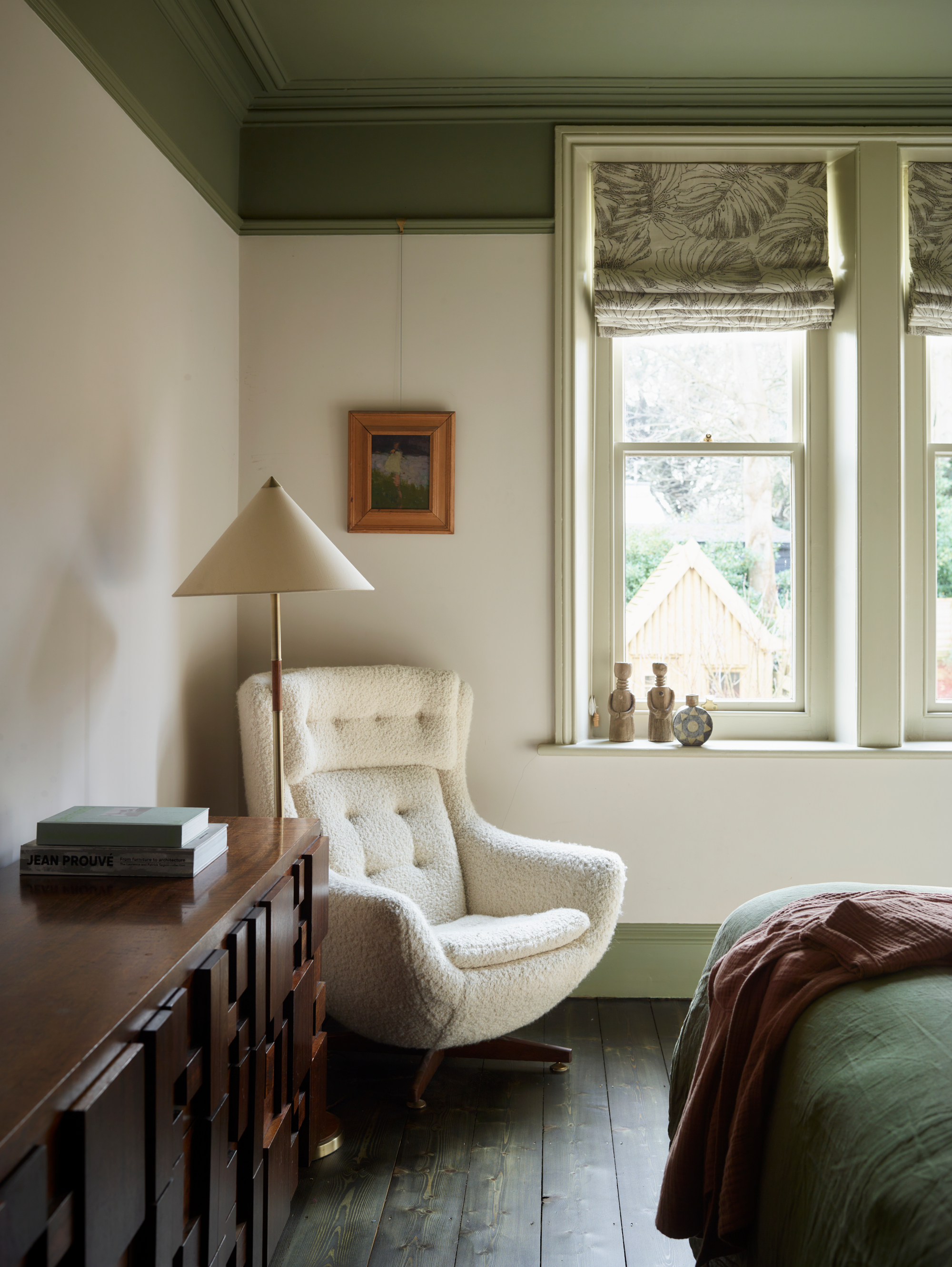 Corner of a bedroom with color-capped green ceiling and white walls, white boucle armchair, cream floor lamp and dark wood sideboard