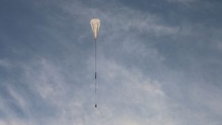 a large white balloon carries a dark, cylindrical payload through a blue sky.