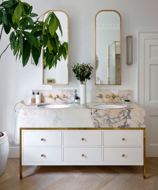 modern bathroom with light wooden flooring in a herringbone style, marble twin basin with white draws beneath, twin arched mirrors above sinks, white painted walls, tall green floor plant in left corner