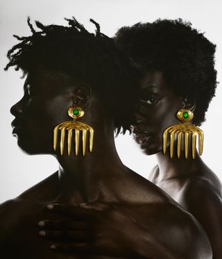 Two models wearing gold earrings with upside down candle forks and a green emerald in the centre.