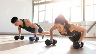 Man and woman side by side performing a push-up using two dumbbells each in a light warehouse gym