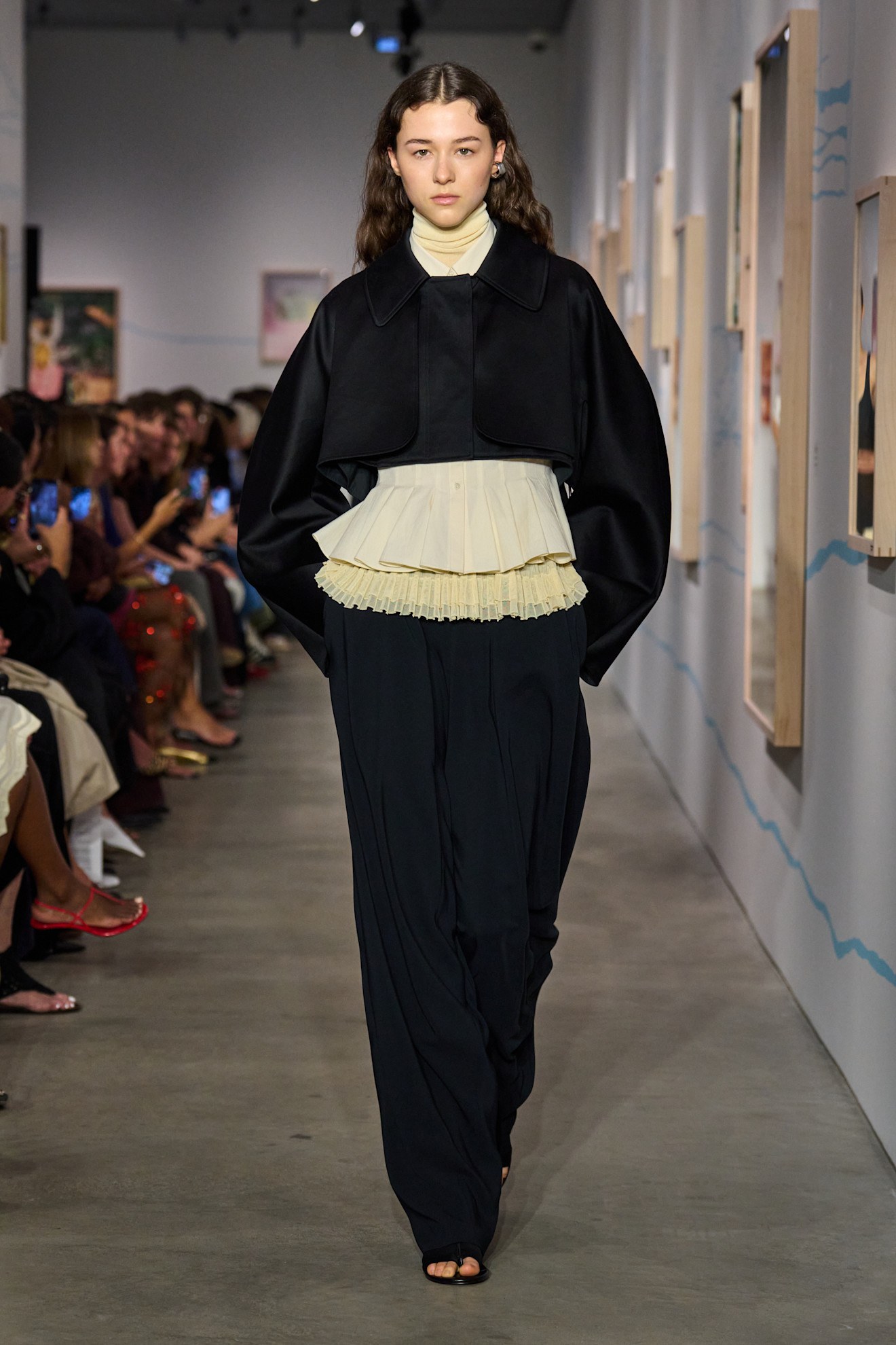 Layering staples for spring 2026 is shown in a photo from Ashlyn's runway show, with a model walking while wearing a butter-yellow turtleneck layered under a pleated blouse and black cropped jacket styled with black wide-leg trousers and black flat sandals