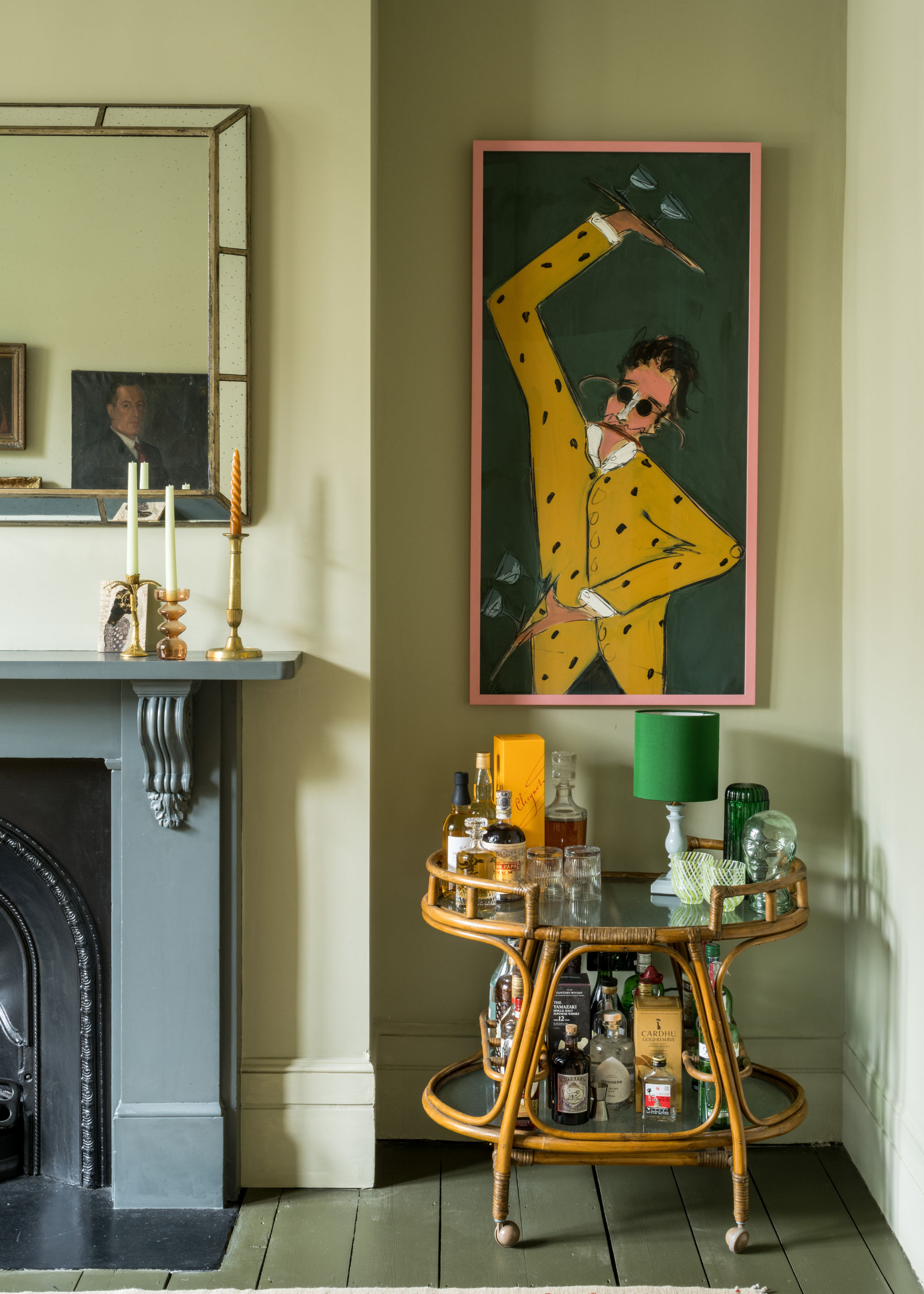 A green living room with a blue mantel place holding tapered candles in candle holders, a large mirror above, and beside a framed painting of a man in a yellow suit above a bar cart with bottles and a green lamp