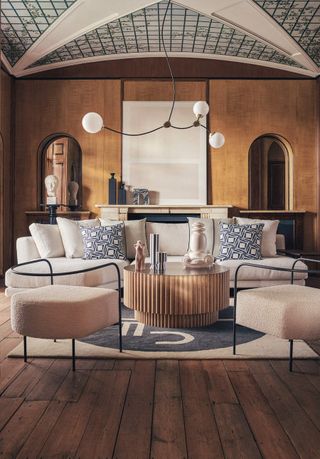 Image of an Art Nouveau-style living room. There are dark wooden floors and the walls are a golden color. There are two arched doorways on either side of the main wall. The ceiling is a curved, geometric pattern. There is a white sofa with two white and black metal accent chairs opposite it.