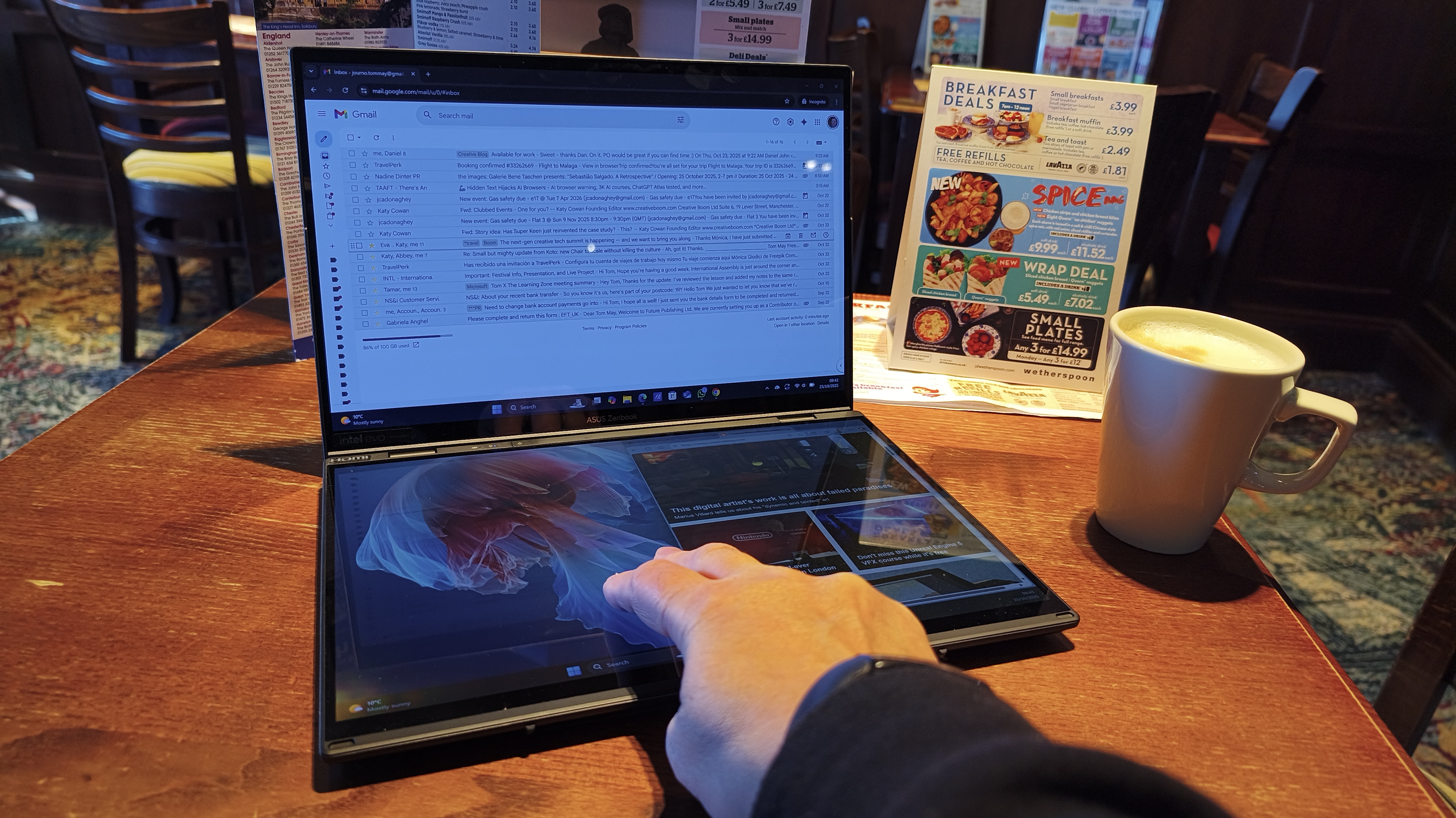 A person's hand is shown touching the bottom display of a dual-screen laptop set up in a folded 'V' shape with a coffee cup and menus on a wooden pub table.