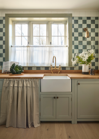 English-style kitchen with sage cabinets, wooden worktops and a curtain-fronted cupboard. The wall is covered in a sage and darker green checkerboard tile print