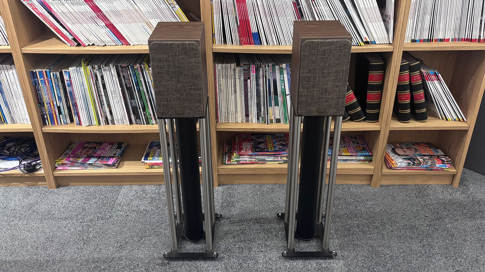 Dali Sonik 1 standmount speakers on stands on grey carpet in front of bookcase, with grilles attached