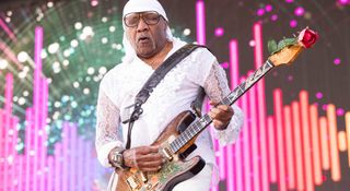 Ernie Isley wears all white and plays his Fender Zeal Stratocaster against a backdrop of pink stage lights. A rose is pinned to its headstock.