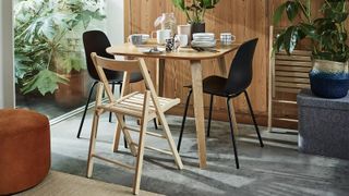 Wooden dining table dressed with tableware for breakfast with one folding wood chair and two black dining chairs in a room with neutral flooring and glass door