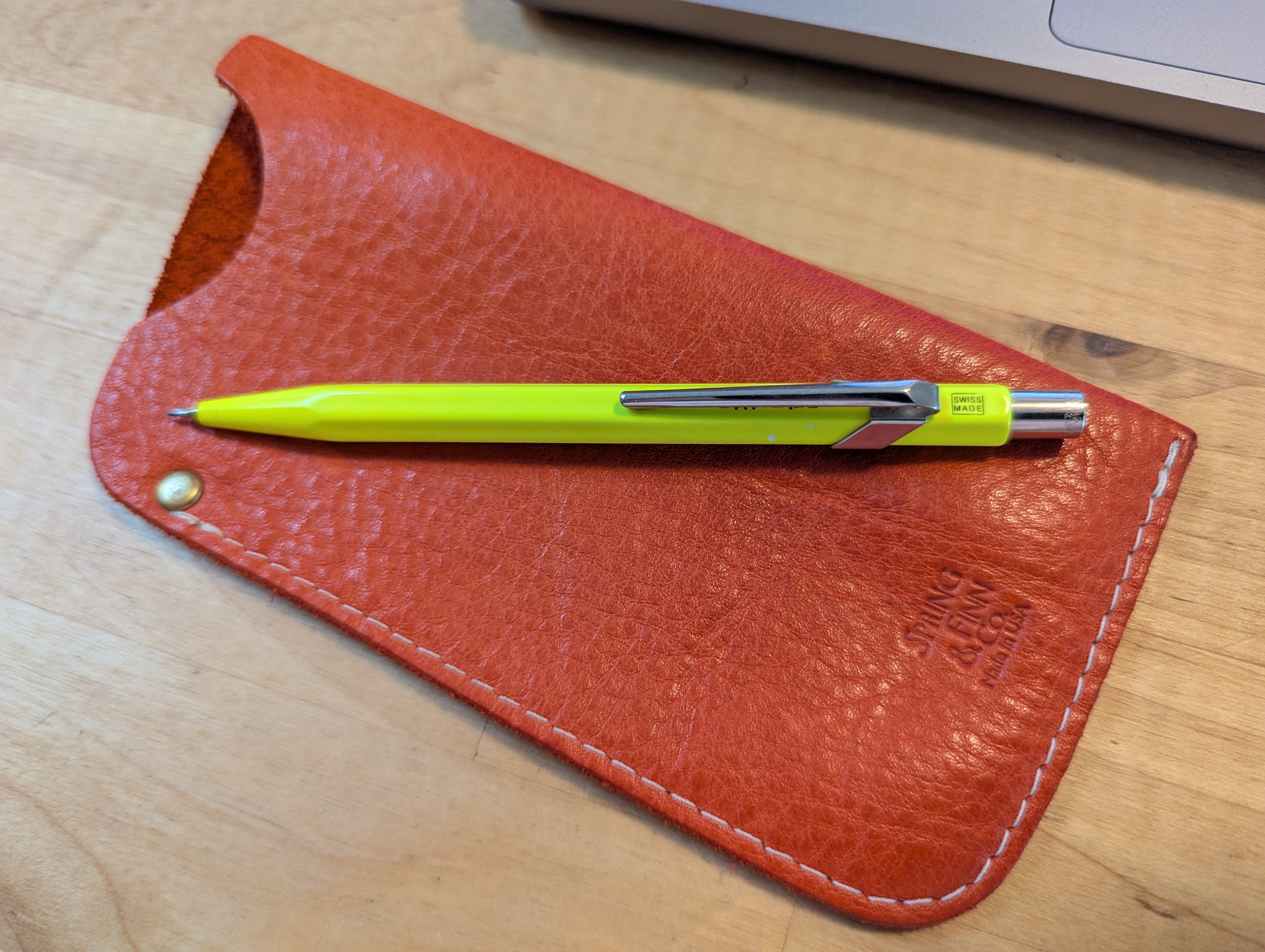 A bright yellow pencil rests on an open, textured orange leather pen sleeve with a small brass button.