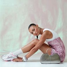 A model sitting on the floor sideways on to the camera with her knees up while wearing a pink tartan dress with white shoulders and Nike trainers in front of a pink and white background